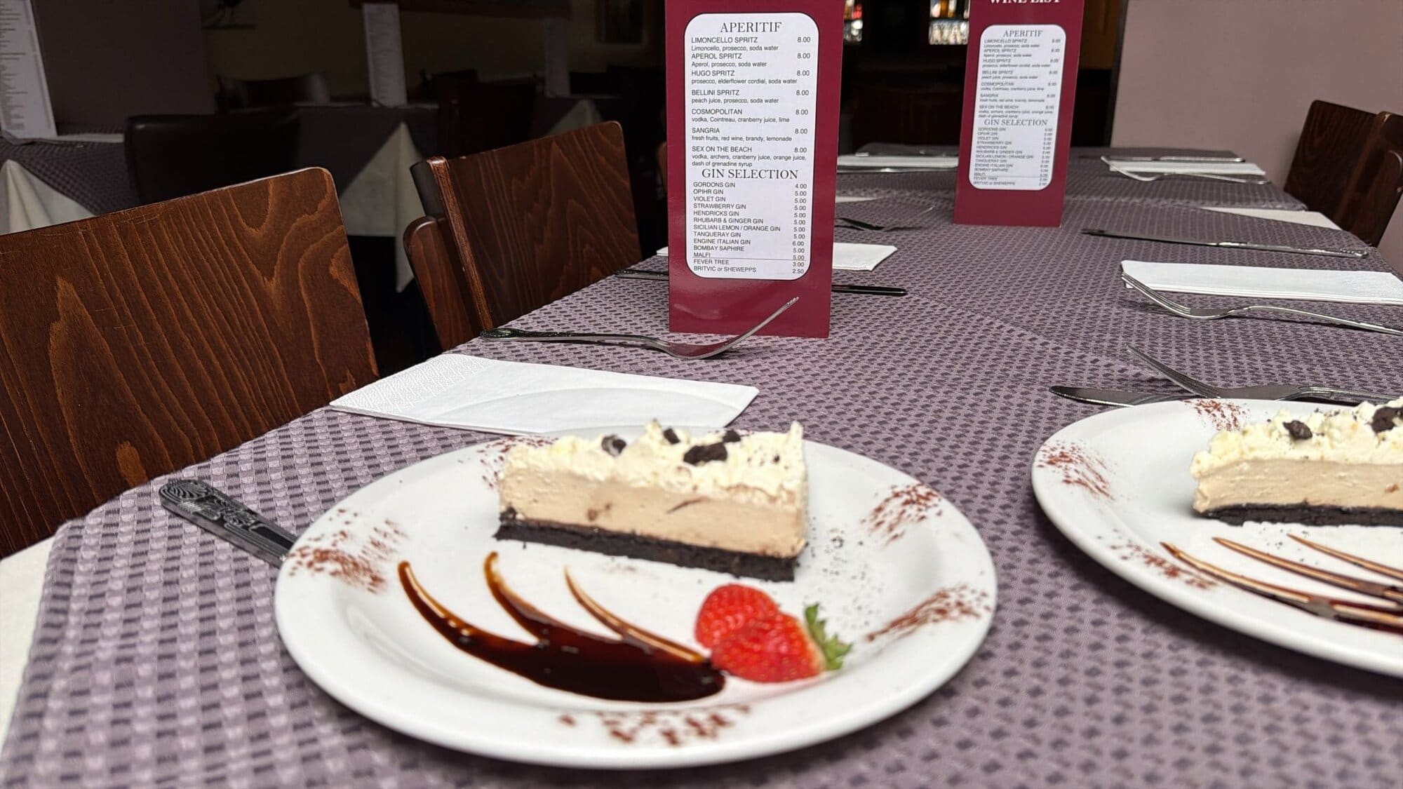 A slice of creamy dessert with chocolate sauce and a strawberry on a laid table in Fabio's dining room, with menus on the tables