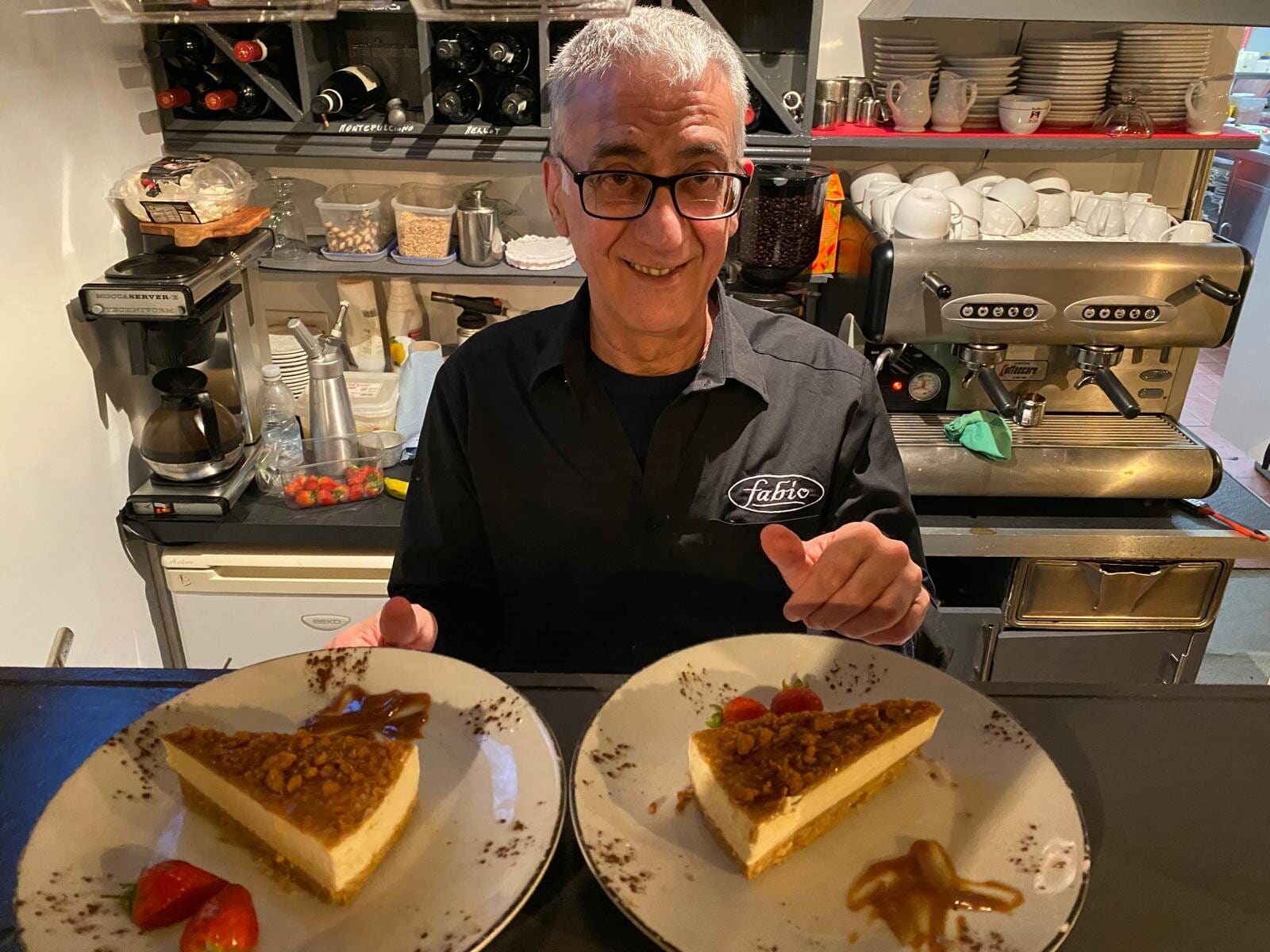 Mario Freti smiling behind the bar at Fabio's in Swindon with two plated slices of cheesecake and strawberries