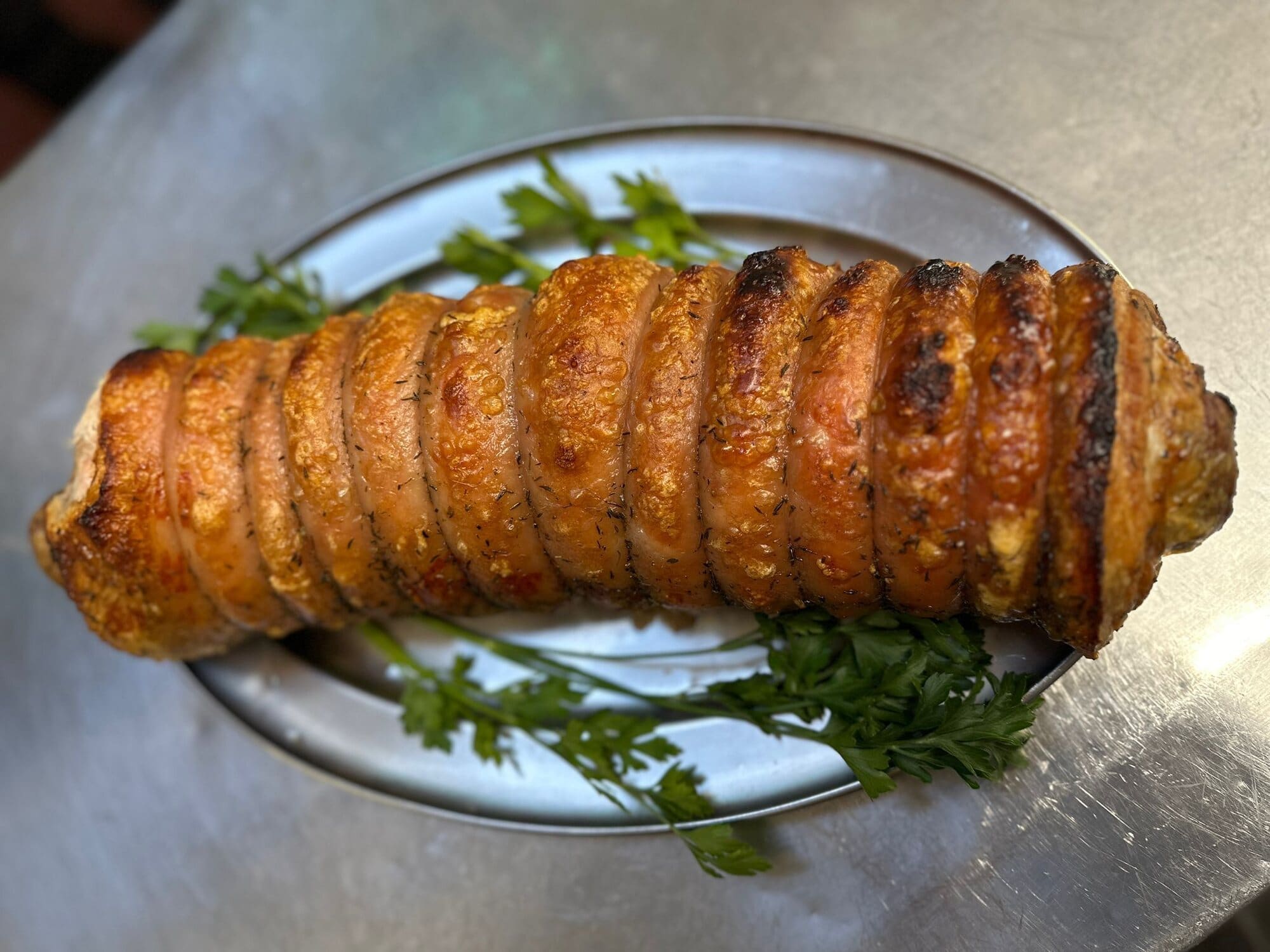 A rolled joint of roast pork with golden crackling and fresh parsley on a silver tray in Fabio's kitchen