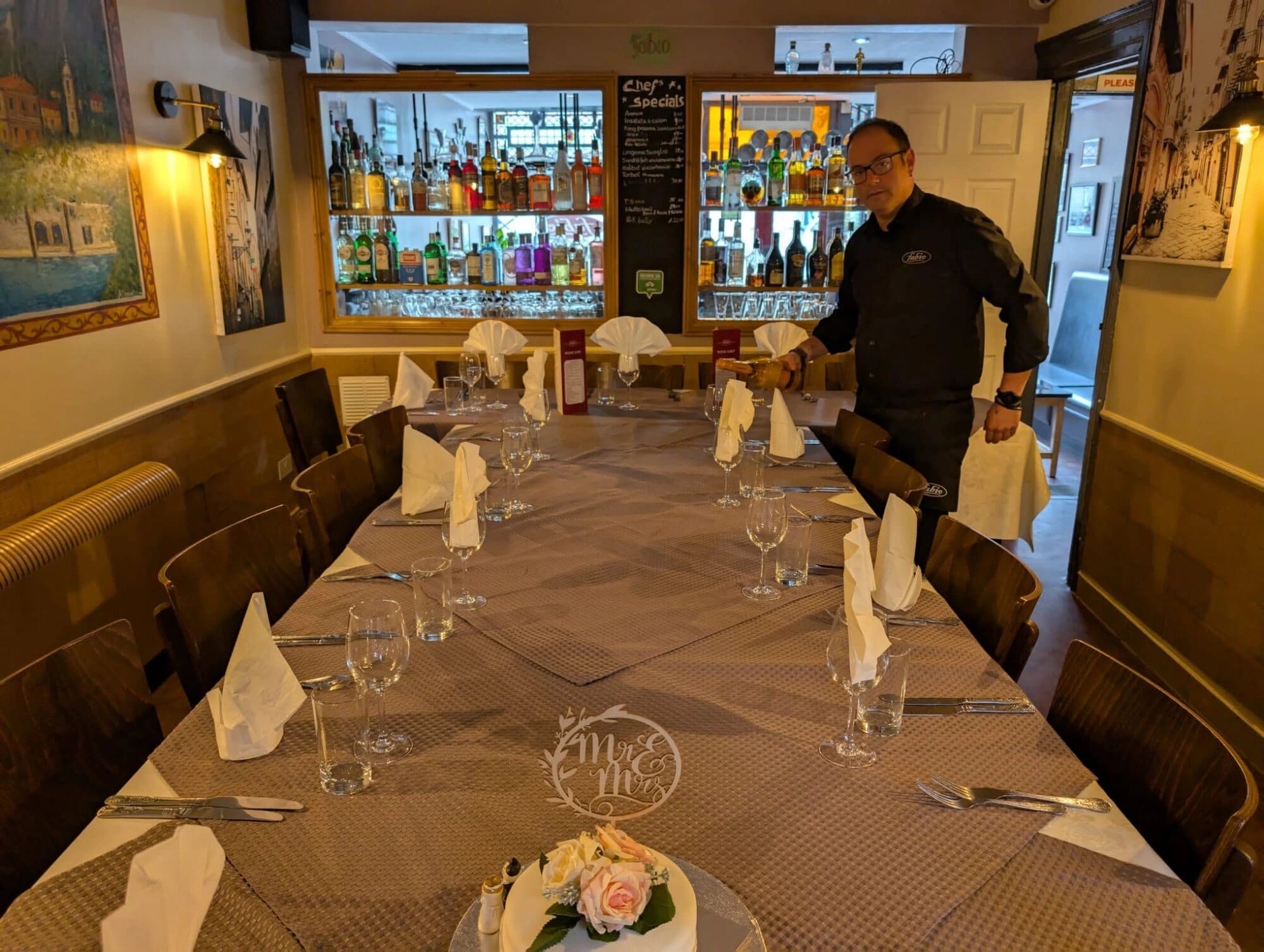 Setting a long table for a wedding party in Fabio's dining room, with the bar and chef's specials board behind