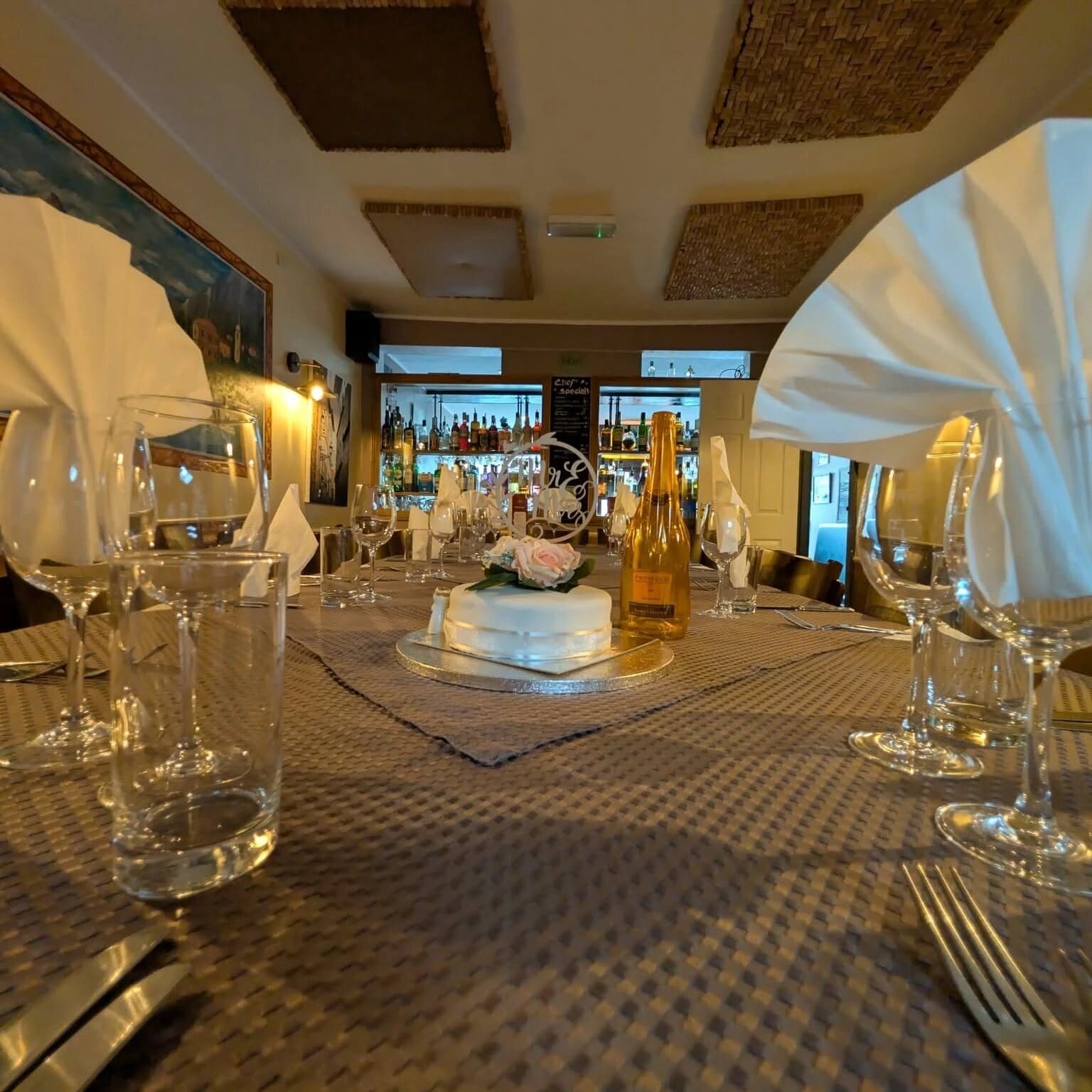 Wedding cake, a bottle of prosecco and laid places on a long table in Fabio's dining room, with the bar behind
